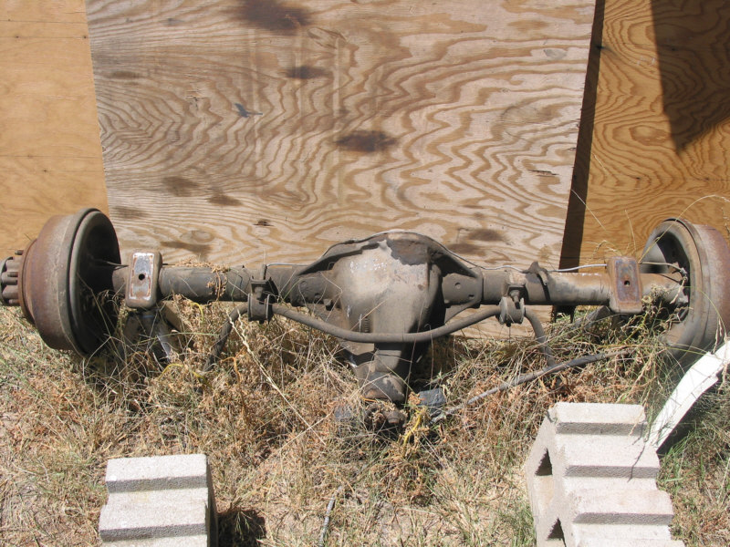Rusty rear axle assembly with drum brakes lying on dry grass against a plywood backdrop