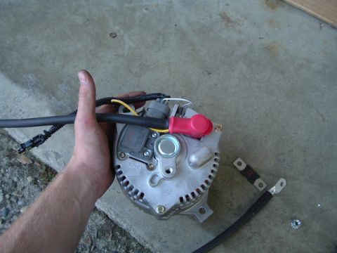 1989 Ford Bronco - A hand holds a car alternator, with thick cables attached, ready for installation.