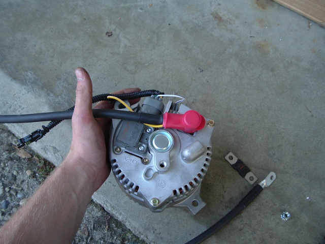 Hand holding a silver alternator with red terminal boot and wiring over concrete