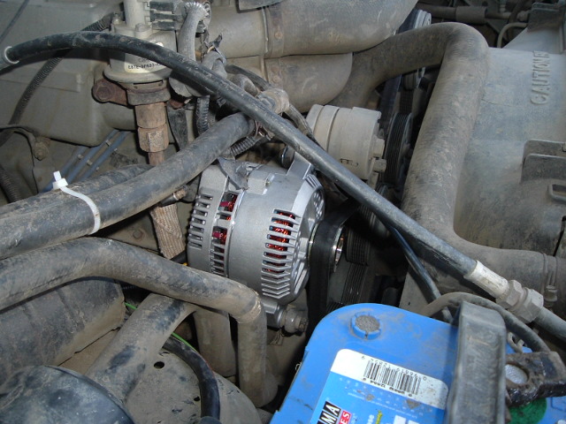 Shiny new alternator installed in a dirty engine bay, serpentine belt attached, blue battery below