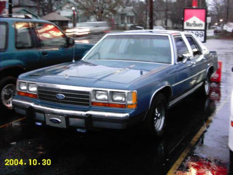 1988 Ford LTD Crown Victoria - Blue Ford Crown Victoria sedan with a white vinyl-look roof parked on wet pavement at a gas station on a rainy day, shot from the front thr…