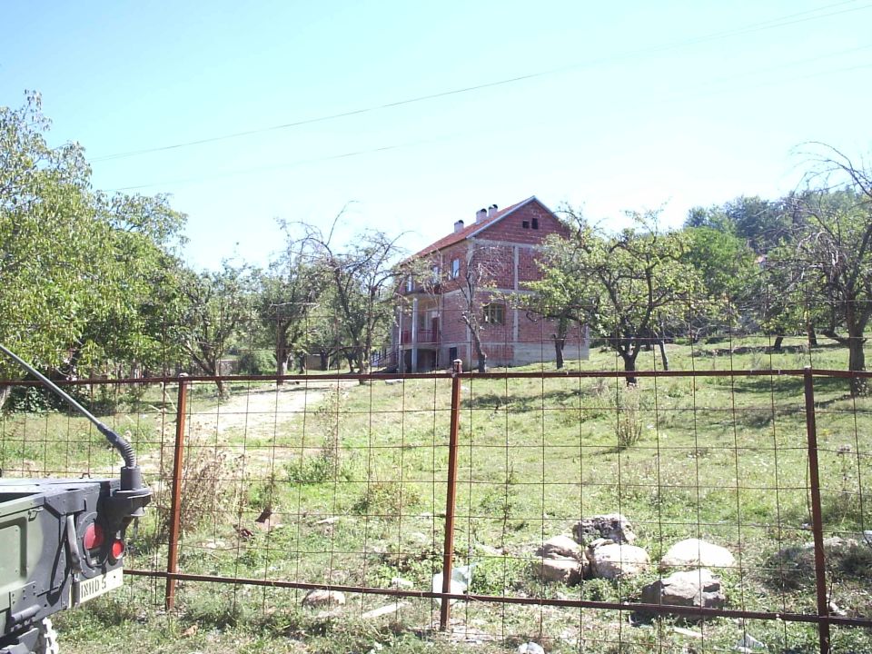 Rear corner of a green military vehicle by a wire fence, with an unfinished brick house in a grassy field behind