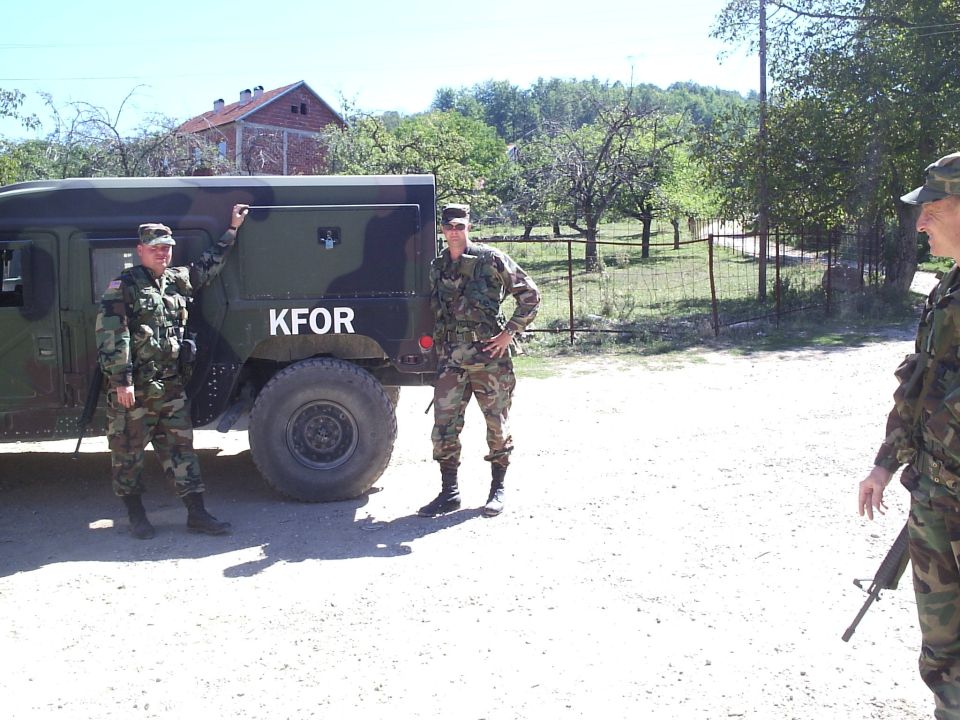 Camouflage military HMMWV marked KFOR on gravel road with three soldiers in woodland camo