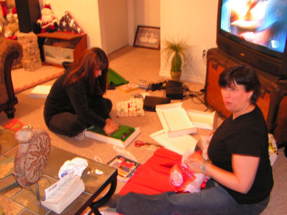 Two women wrapping gifts on a living room floor near a TV and Christmas decorations