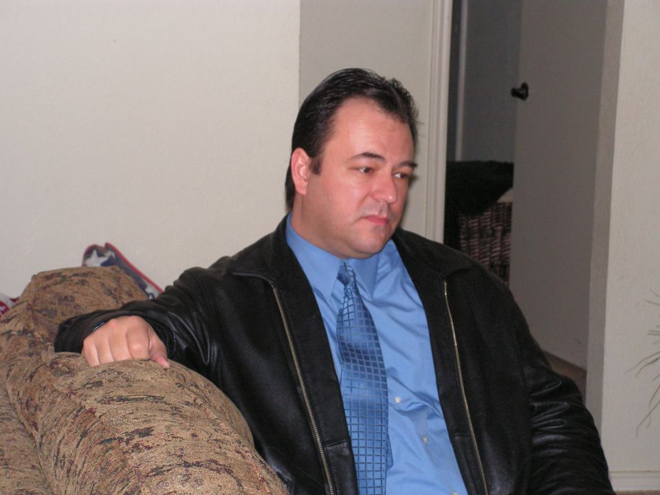 Man in black leather jacket and blue shirt with tie seated against a sofa