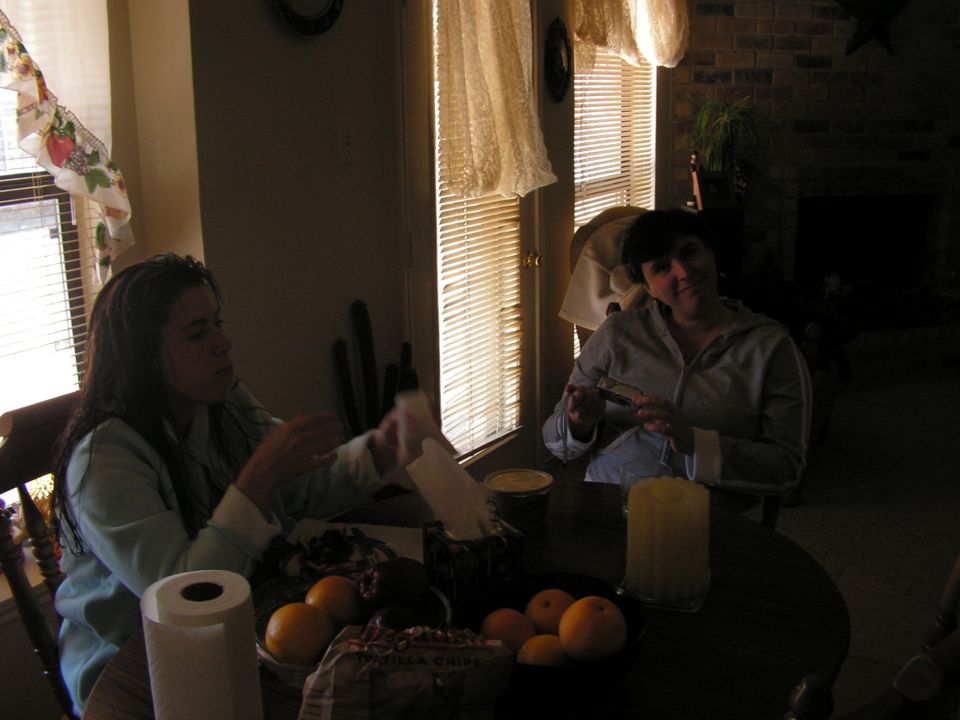 Two women sit at a table with fruit and paper towels; one woman holds a small object