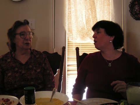 1978 Ford Bronco - Two women are seated at a dining table, engaged in conversation near a window with blinds and lace curtains.