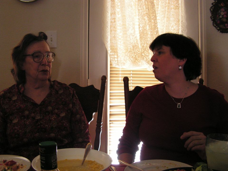 Two women sit at a table, one wearing glasses and a patterned shirt, the other a maroon sweater