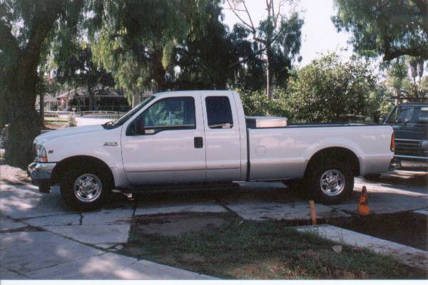 Side view of a white Ford Super Duty extended-cab long-bed pickup with bed toolbox, parked in a shaded driveway