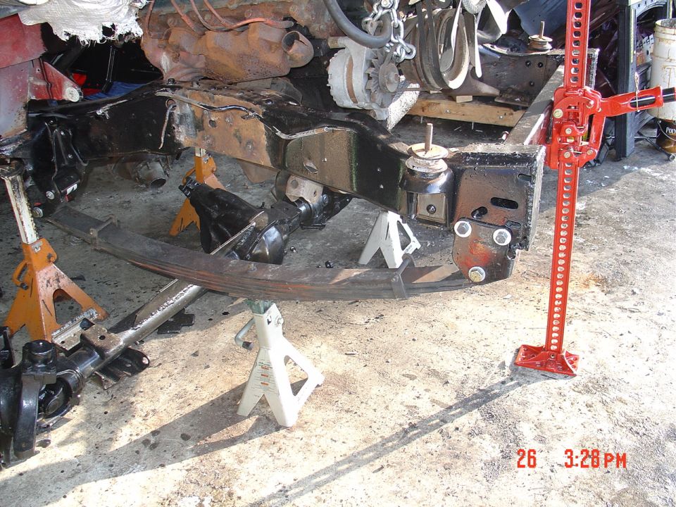 Stripped pickup frame on jack stands with engine, leaf spring, removed axle, and red Hi-Lift jack in a garage