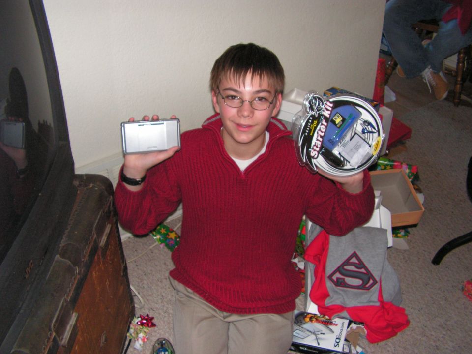 Young boy in glasses and a red sweater holds a silver electronic device and a circular gift package. Presents are scattered around him