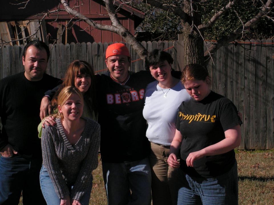 Group of six people posing for a photo in front of a wooden fence and tree