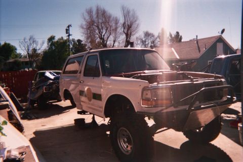 1990 Ford Bronco - white full-size Ford Bronco stripped of doors with a tube front end and lifted suspension, parked in a residential driveway.