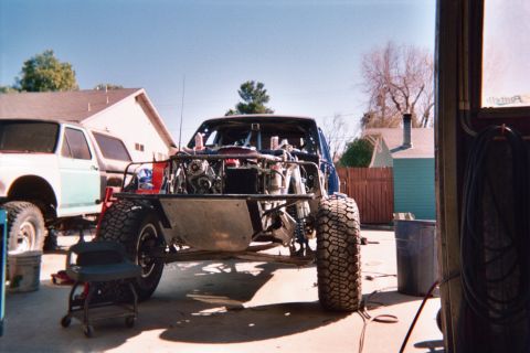 1990 Ford Bronco - Front view of a tube-frame off-road truck build with exposed engine and large mud tires, parked beside a lifted SUV in a driveway.