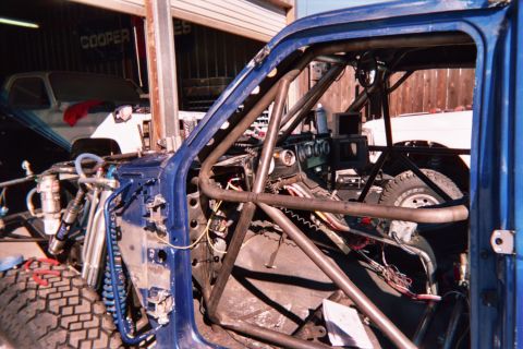 1990 Ford Bronco - blue truck cab fitted with a welded tubular roll cage and exposed wiring during an off-road vehicle build in a shop yard.