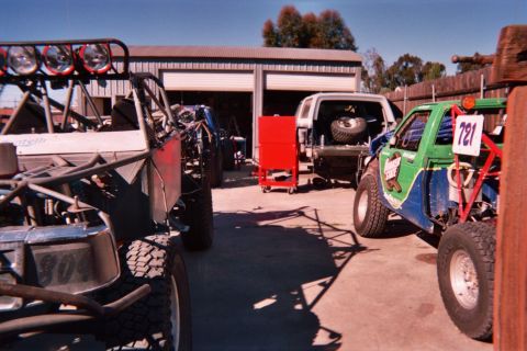 1990 Ford Bronco - cluster of off-road race buggies and trucks parked outside a shed, including a green car numbered 721 and a light-bar rig.