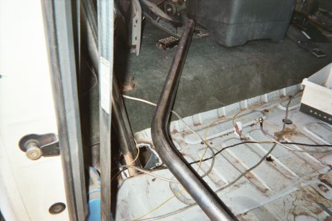 1990 Ford Bronco - Interior corner of a stripped vehicle showing a bent tubular cage bar running over worn carpet and the bare floor pan.