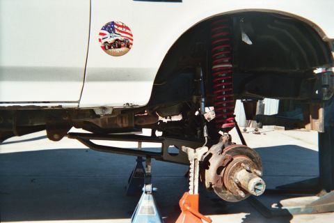 1990 Ford Bronco - white truck raised on jack stands with a red coil-over shock, brake drum hub and tube lower arm exposed at the wheel well.