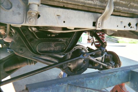 1990 Ford Bronco - Underbody view of a solid front axle with differential housing, tie rod and a red coil-over shock on a raised vehicle.