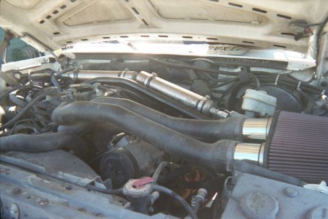 1990 Ford Bronco - Engine bay close-up showing the exhaust manifold, a red cone air filter intake and brake master cylinder under an open hood.