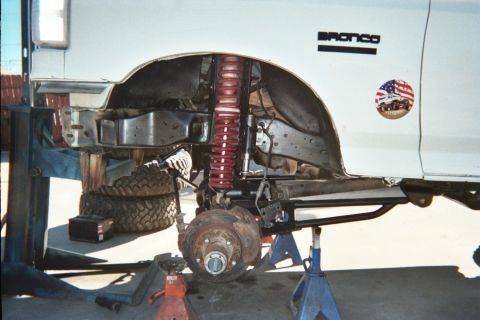 1990 Ford Bronco - white Ford Bronco raised on jack stands, BRONCO badge visible, with a red coil-over shock and exposed front brake hub.