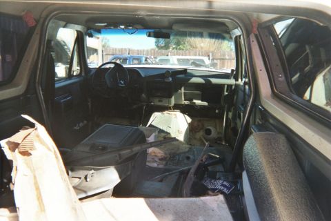 1990 Ford Bronco - Gutted SUV interior seen from the cargo area, showing bare floor pan, dashboard and steering wheel with a parking lot visible ahead.