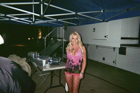 1990 Ford Bronco - smiling blonde woman in a pink top and fishnet tights stands under a dark canopy beside a folding table and white trailer at night.