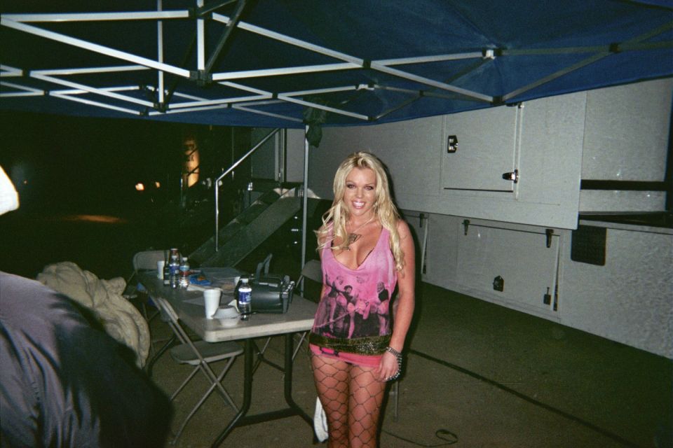 Smiling blonde woman in a pink top and fishnets standing under a canopy near a white trailer at night