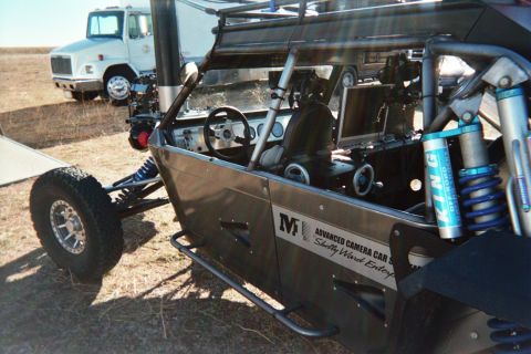 1990 Ford Bronco - black open tube-chassis vehicle marked Advanced Camera Car parked on dry grass with a flatbed truck behind.
