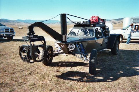 1990 Ford Bronco - black tube-framed vehicle with a long boom arm and red gear bag on its roof rack stands in an open grassy field.