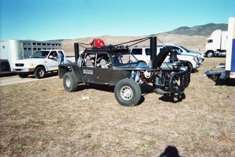1990 Ford Bronco - black open-frame off-road vehicle rigged with a rear camera boom sits on a dirt lot beside pickups and semi trailers.