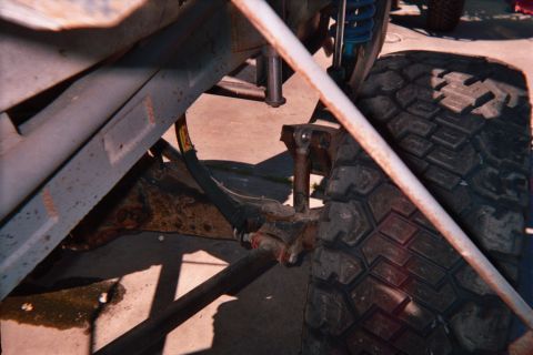 1990 Ford Bronco - Low underside shot showing a rusty frame, leaf spring and steering linkage beside a knobby off-road tire on concrete.