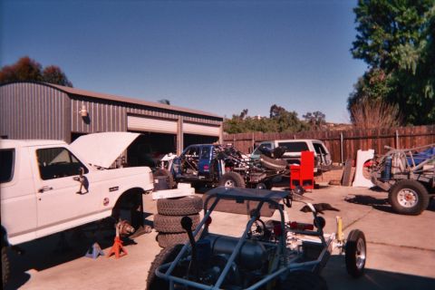 1990 Ford Bronco - driveway workshop scene with a white pickup hood-up, tube-chassis buggies, stacked tires and a sand rail under clear sky.