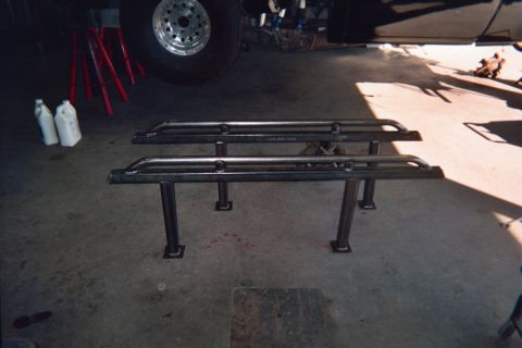 1990 Ford Bronco - Two black welded tubular steel rack frames sit on a garage floor near red brackets and a chrome wheel.
