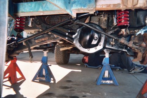 1990 Ford Bronco - truck raised on blue and red jack stands revealing its rear axle housing, driveshaft and red coil springs.