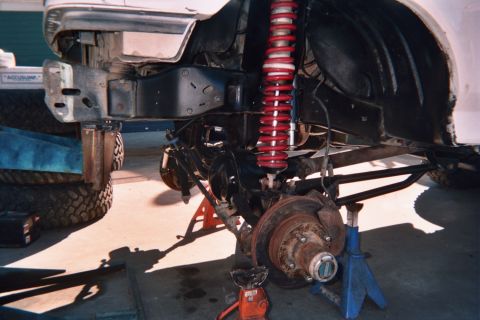 1990 Ford Bronco - Front corner of a white truck on jack stands showing a red coilover shock, brake rotor and disassembled suspension.