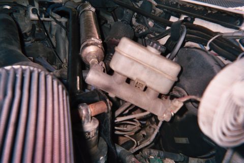 1990 Ford Bronco - Engine-bay view of a translucent brake master cylinder reservoir beside a red conical air filter and tangled hoses.