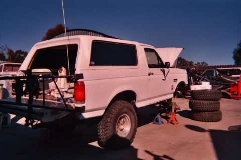 1990 Ford Bronco - white lifted Ford Bronco on knobby tires with its rear hardtop window raised, parked on a dirt lot near stacked tires.