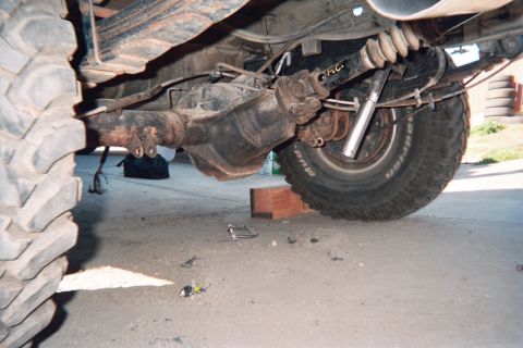 1990 Ford Bronco - Under a lifted truck showing the differential housing, driveshaft and an off-road tire resting on a wood block.