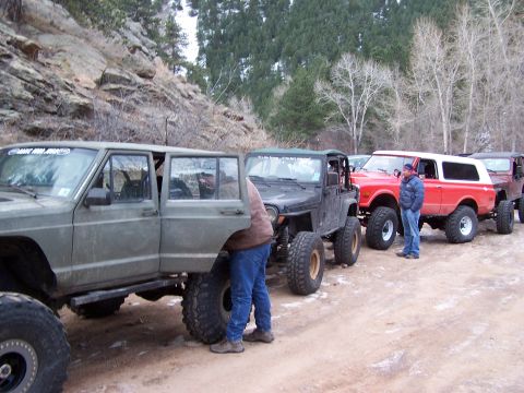 1985 Ford Bronco - Several modified off-road vehicles are parked in a line on a dirt road, with a rocky hillside in the background.