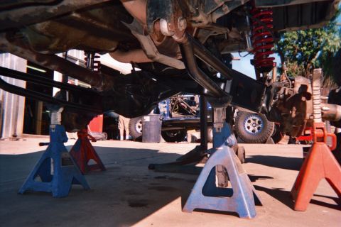 1990 Ford Bronco - Low-angle underbody view of a lifted truck supported on blue and red jack stands, showing a red coil-over shock and exhaust during a build.