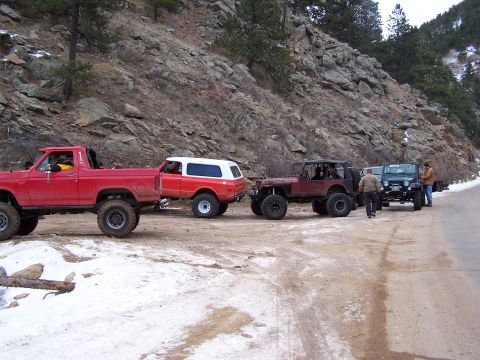 1985 Ford Bronco - A group of off-road vehicles are parked on a dirt road in a rocky, wooded area with snow on the ground.