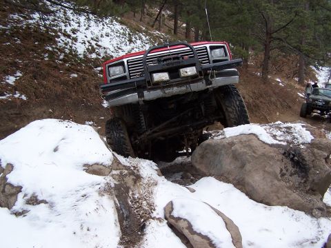 1985 Ford Bronco - A red off-road truck maneuvers through a rocky, snow-covered trail.