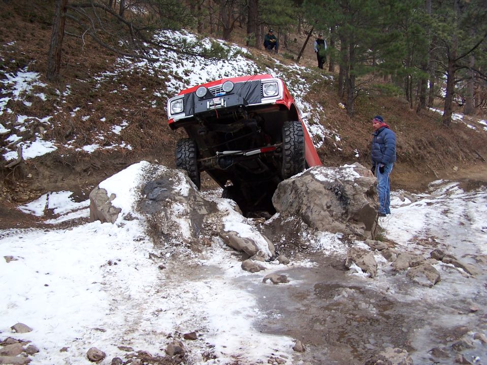 Red off-road vehicle with large tires stuck between rocks on a snowy trail. Two people watch from a hillside