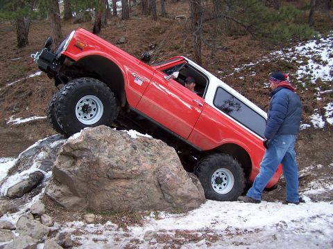 1985 Ford Bronco - A man stands by as a red SUV navigates a difficult rocky obstacle on a winter trail.