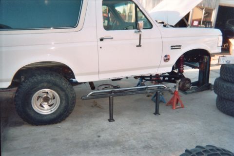 1990 Ford Bronco - white Ford Bronco sits on jack stands inside a garage with one front wheel removed exposing the suspension.