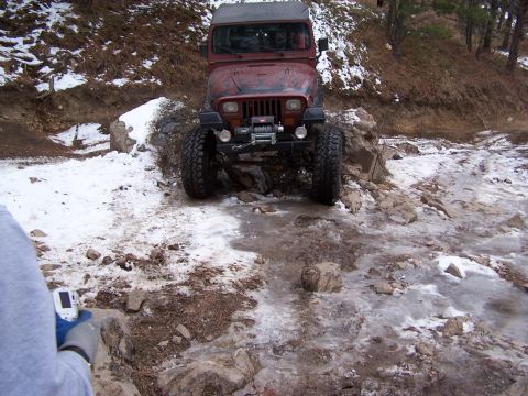 1985 Ford Bronco - A red Jeep Wrangler navigates a challenging off-road trail with icy and snowy terrain.