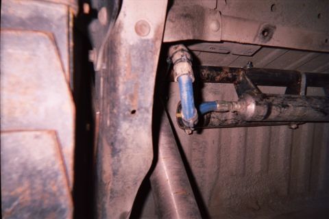 1990 Ford Bronco - Tight undercarriage shot of a vehicle frame rail and crossmember with a blue-painted plumbing fitting attached.