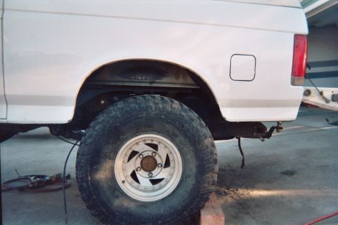 1990 Ford Bronco - Rear quarter of a white lifted truck showing a mud-terrain tire mounted on a white five-spoke alloy wheel.