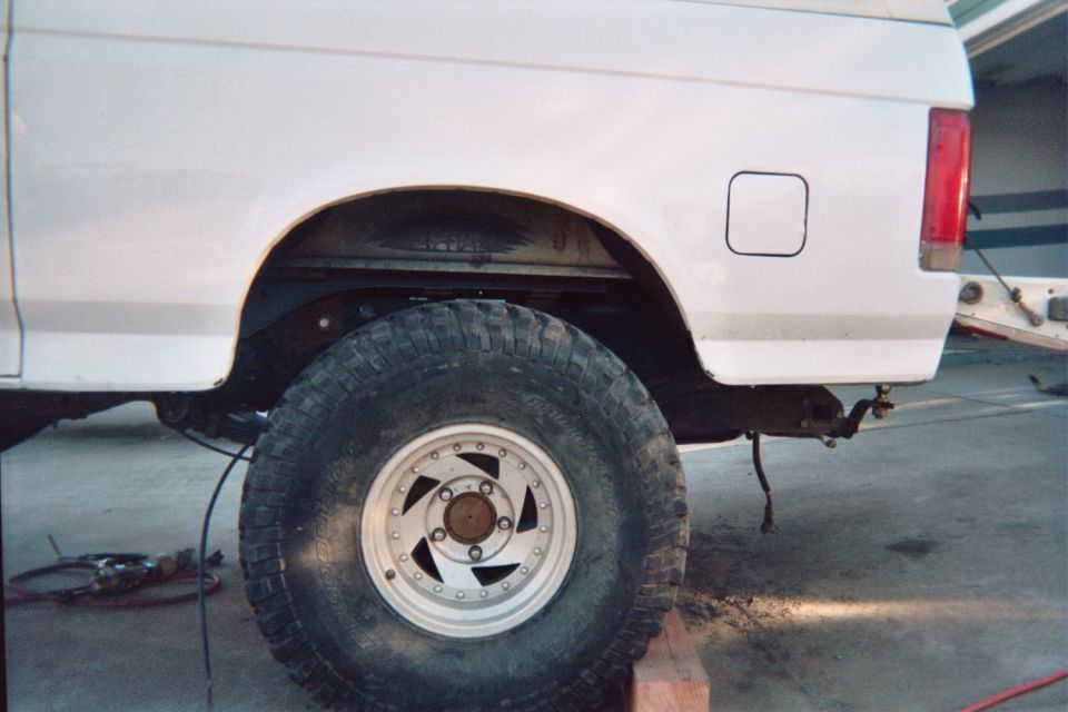 Rear quarter of a white truck or SUV with a mud tire on a white five-spoke wheel, in a garage